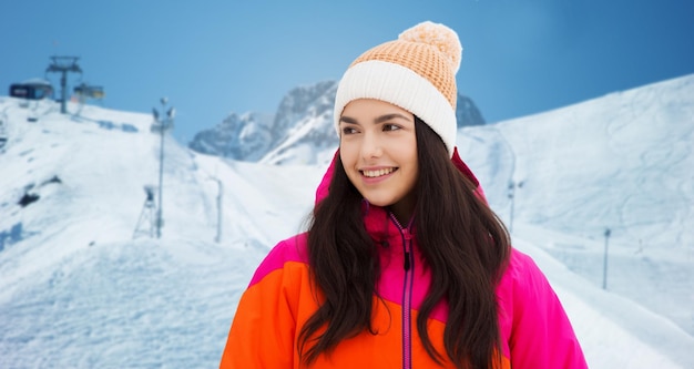 Winter-, Freizeit-, Kleidungs- und Personenkonzept - glückliche junge Frau oder Teenager-Mädchen in Winterkleidung über Abfahrtslauf und Berghintergrund