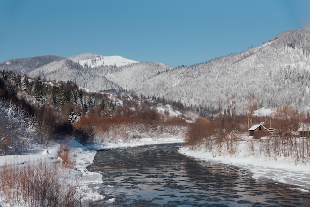 Winter, frostiger Tag in einem Dorf zwischen den schneebedeckten Bergen am Ufer des Flusses.