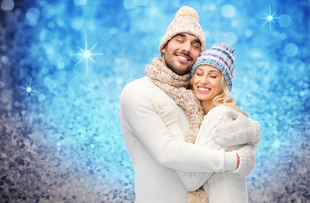 Winter-, Mode-, Paar-, Weihnachts- und Personenkonzept - lächelnder Mann und Frau in Hüten und Schal, die sich über blauem Glitzer oder hellem Hintergrund umarmen