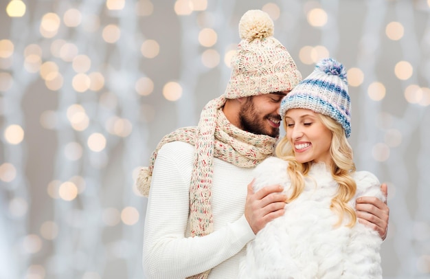 Winter-, Mode-, Paar-, Weihnachts- und Personenkonzept - lächelnder Mann und Frau in Hüten und Schal, die sich über Feiertage umarmen, beleuchtet den Hintergrund