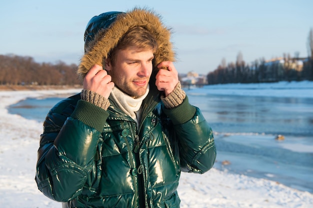 Winter-Outfit. Hipster-Wintermode-Outfit. Kerl trägt Jacke mit Kapuze an frostigen Wintertagen. Mann bärtiger Stand warme Jacke verschneite Natur Hintergrund. Windabweisende Kleidung. Winter stilvolle Herrenmode.