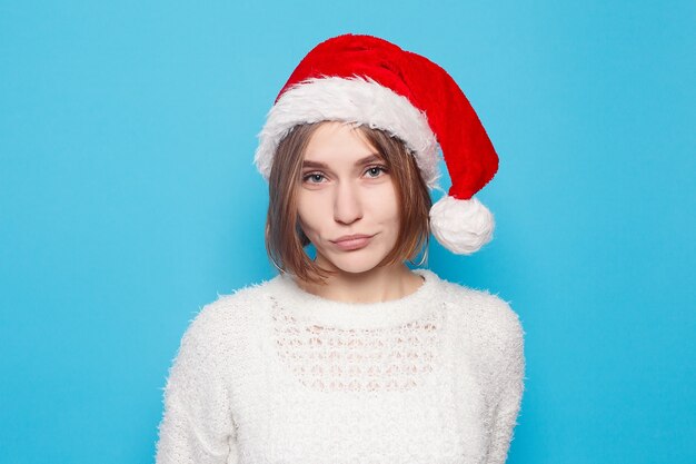 Winter, Weihnachten, Leute, Schönheitskonzept - hübscher blonder tragender Weihnachtshut auf hellblauem Hintergrund. Porträt des jungen schönen netten fröhlichen Mädchens, das Kamera über weißem Hintergrund betrachtet.