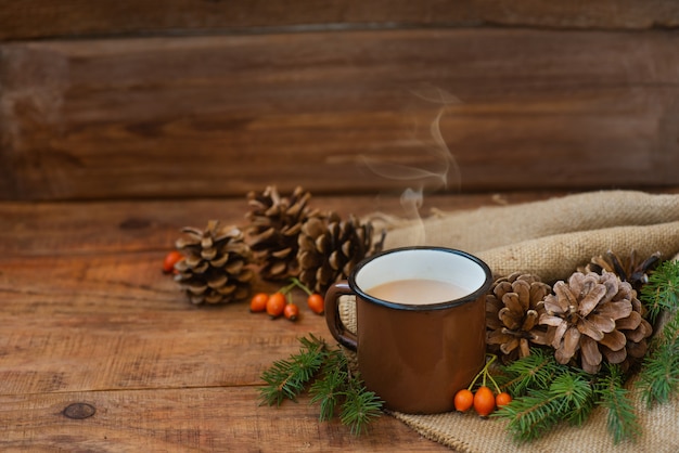 Winter, Weihnachtshintergrund im rustikalen Stil. Ein Vintage-Metallbecher mit heißem Milchtee steht auf einer Tischdecke, auf einer Holzoberfläche zwischen Tannenzapfen, Fichtenzweigen und Hagebutten. Platz kopieren, flach legen