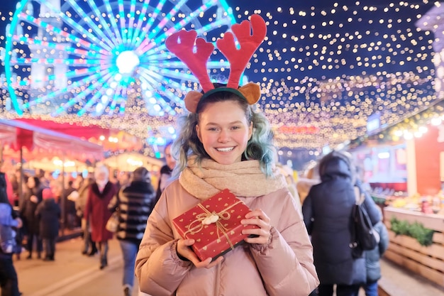 Winter-Weihnachtsporträt des glücklichen Jugendlichen mit Geschenkbox am Feiertags-Stadtmarkt im Freien. Lichter, helle Girlanden der Abendstadt, Riesenradhintergrund