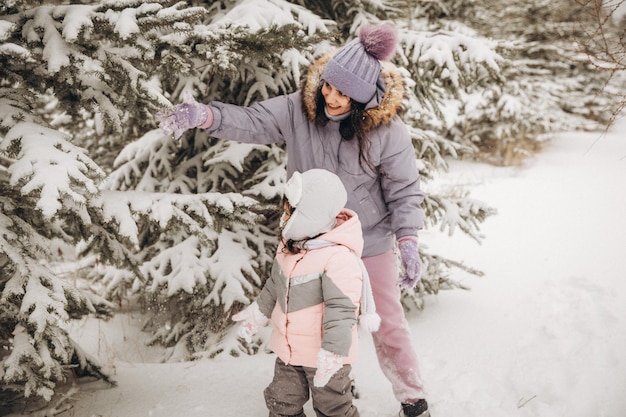 Wintererholung im Freien. Glückliche Mutter schüttelt Schnee von einem Ast auf einem Kind ab, das zwischen schneebedeckten Tannen steht