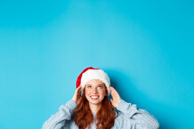 Winterferien und Heiligabend Konzept. Kopf des niedlichen rothaarigen Mädchens im Weihnachtsmannhut, erscheinen von unten und schaut nach oben zum Kopienraum, starrendes Logo, stehend über blauem Hintergrund.