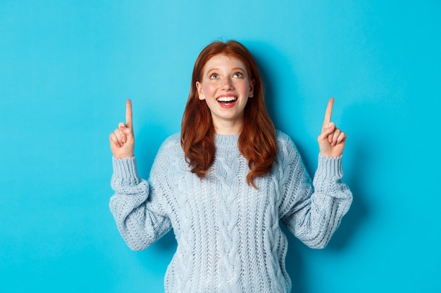 Winterferien und Menschenkonzept. Schönes Teenager-Mädchen mit roten Haaren, Pullover tragend, mit den Fingern nach oben auf das Logo zeigend und lächelnd, Werbung zeigend, blauer Hintergrund