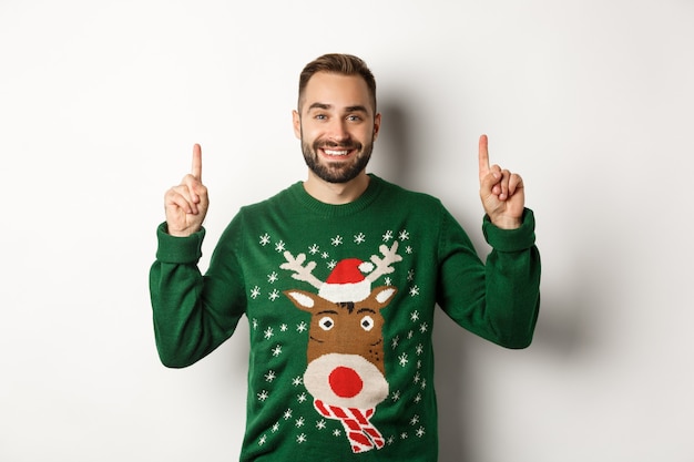 Winterferien und Weihnachten. Schöner bärtiger Mann im grünen Pullover, der mit den Fingern nach oben zeigt und lächelt, Werbung zeigt, weißer Hintergrund