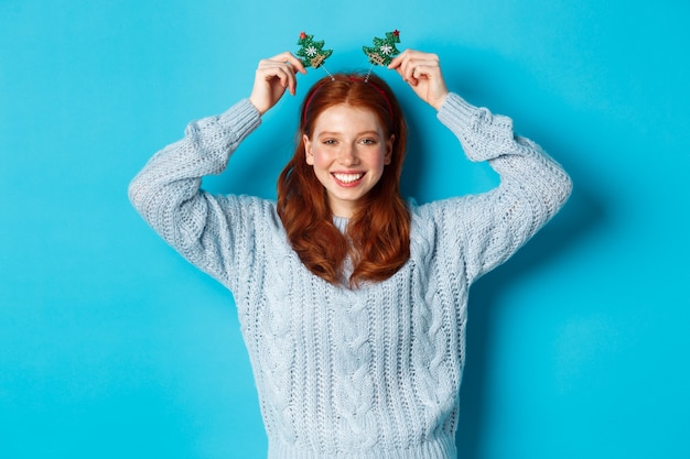 Winterferien und Weihnachtsverkaufskonzept. Schönes weibliches Modell der Rothaarigen, die Neujahr feiern, lustiges Party-Stirnband und Pullover tragen und in die Kamera lächeln.