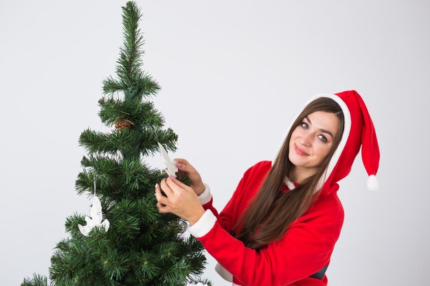 Winterferien-, Weihnachts- und Personenkonzept - Porträt der schönen jungen Frau in der Weihnachtsmütze, die Weihnachtsbaum zu Hause verziert