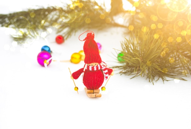 Winterfoto mit Weihnachtsschmuck für Kiefer. Gestricktes Schneemann-Spielzeugmädchen, das auf dem Ski im Wald steht. Frohe Feiertage verziert Postkarte. Verschneites Wetter.