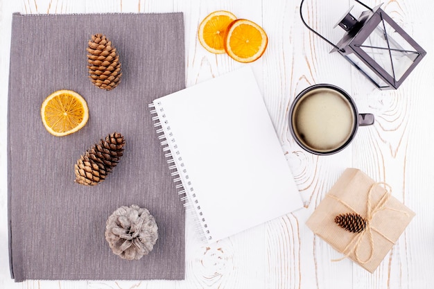 Winterkomposition mit leerem Tagebuch Tasse Kaffee Tannenzapfen Laterne gebackene Orangenscheiben Geschenk verpackt in Kraftpapier auf rustikalem weiß getünchten Holzhintergrund Flacher Kopierraum