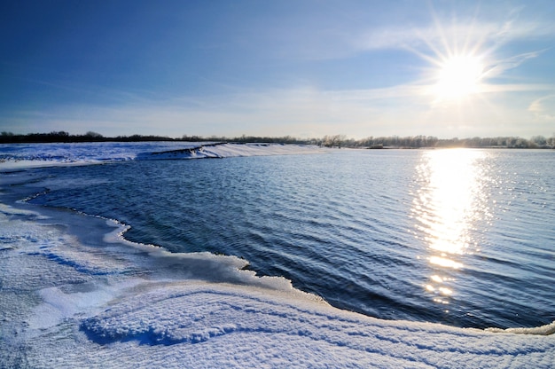 Winterlandschaft, blauer Himmel und strahlendes Sonnenlicht, schneebedecktes Seeufer, Bäume wachsen am Horizont