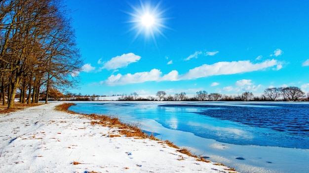 Winterlandschaft mit der Sonne im blauen Himmel über dem Fluss und den Bäumen