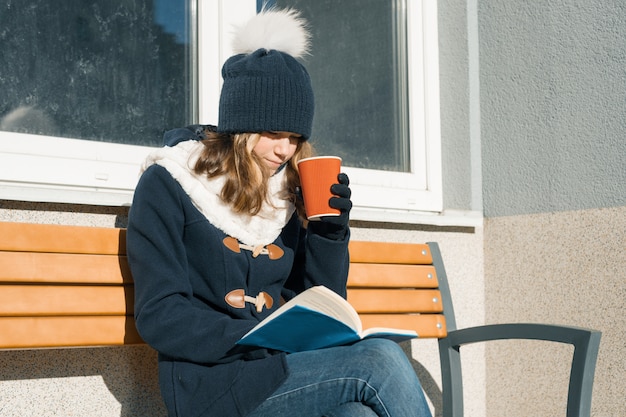 Winterporträt des mädchens des jungen jugendlich mit schale des heißen getränks und des buches.