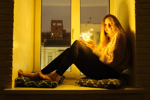 Winterporträt einer jungen schönen Frau, die in der Nähe eines Fensters auf der Fensterbank sitzt, nächtlicher festlicher Stadthintergrund, funkelnde Girlandenlichter in den Händen eines Mädchens