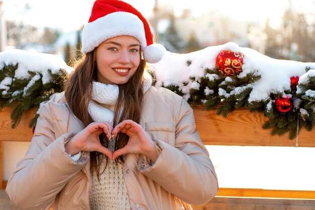 Winterporträt junge erwachsene schöne Frau in Weihnachtsmütze mit Herzform durch Finger Weihnachtsstimmung Schneiendes Winterschönheitsmodekonzept