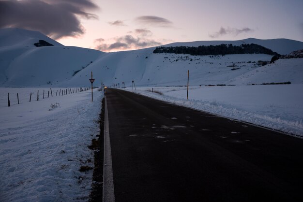 Winterstraße in der Abenddämmerung nach Sonnenuntergang, umgeben von schneebedeckten Bergen