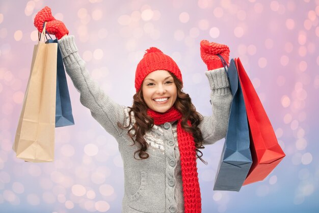 Winterurlaub, Ausverkauf, Weihnachten und Menschenkonzept - lächelnde junge Frau mit Hut und Fäustlingen mit Einkaufstüten über Rosenquarz und heiteren Lichtern im Hintergrund