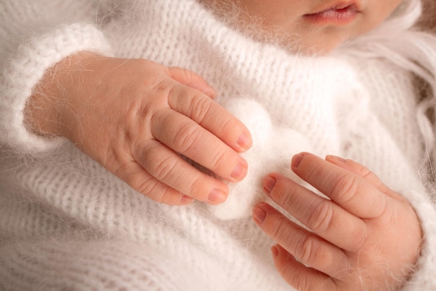 Winzige Hand eines neugeborenen Babys. Weiche Hände von Neugeborenen in weißen Kleidern halten weiße Wollherzen. Finger eines neugeborenen Babys hautnah. Studio-Makrofoto. Glück einer Frau. Fotografie, Konzept.