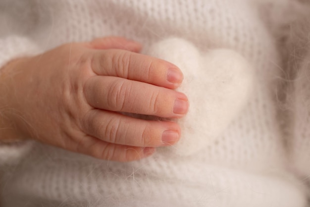 Winzige Hand eines neugeborenen Babys. Weiche Hände von Neugeborenen in weißen Kleidern halten weiße Wollherzen. Finger eines neugeborenen Babys hautnah. Studio-Makrofoto. Glück einer Frau. Fotografie, Konzept.