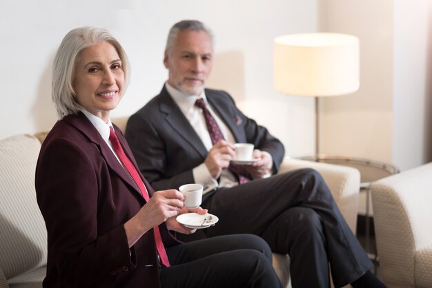 Wir genießen Kaffee. Fröhlich entzückte alte Geschäftsfrau, die lächelt und im Büro sitzt, während sie mit ihrer Kollegin einen Kaffee trinkt und Glück ausdrückt