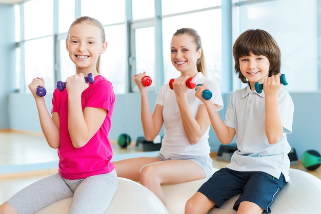 Wir halten unseren Körper fit. Fröhliche Mutter und zwei Kinder trainieren mit Hanteln im Fitnessstudio, während sie zusammen auf den Fitnessbällen sitzen sitting