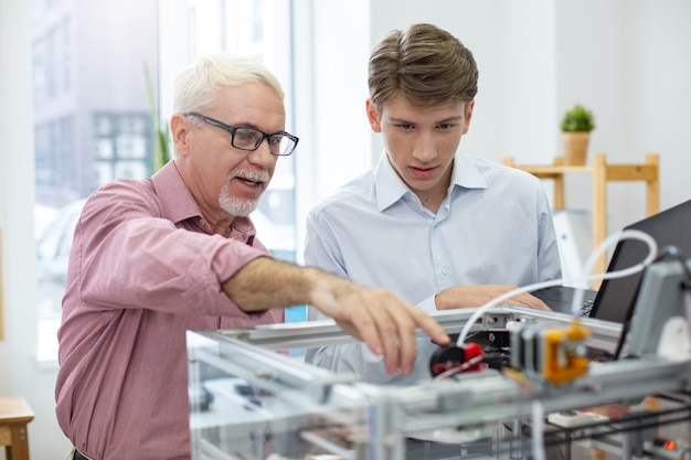 Wissen weitergeben. Erfahrener leitender Ingenieur, der seinen jungen Praktikanten über 3D-Drucker unterrichtet und dabei auf die wichtigen Teile des Mechanismus hinweist