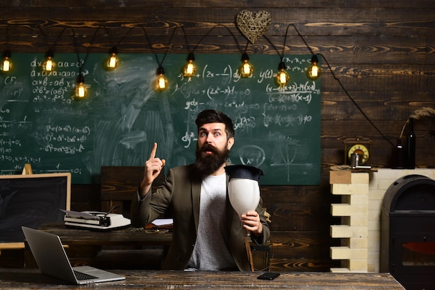 Wissenschaftler Hipster mit Glühbirne auf Tafel Bärtiger Mann hält Glühbirne im Klassenzimmer Geschäftsmann im Anzug an der Schulbank Lehrer mit langem Bart auf genialem Gesicht Habe Idee Erleuchtung und Erfindung
