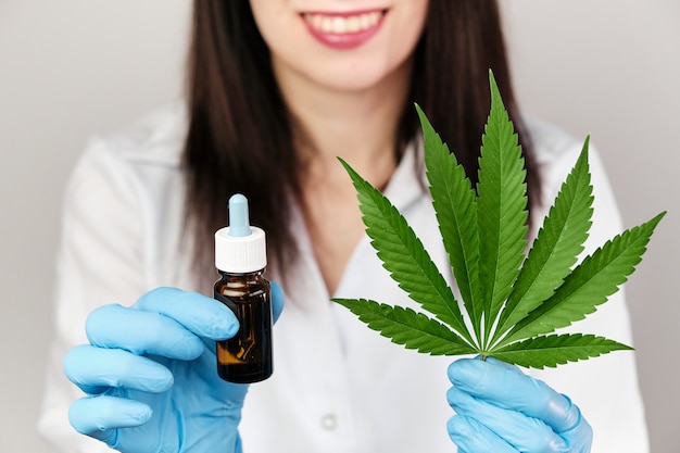 Wissenschaftler oder Arzt in blauen Gummihandschuhen und Laborkittel mit CBD-Hanföl in einer Durchstechflasche und einem Cannabisblatt. Alternativmedizin oder Apothekenkonzept im Gesundheitswesen. Medizinische Marihuana-Pflanze.