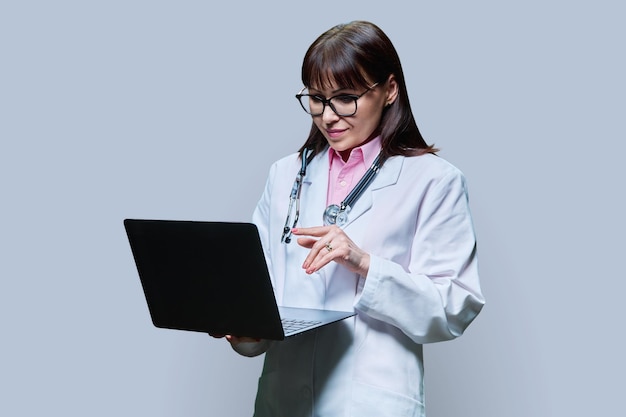 Wman médico usando laptop olhando para a câmera em fundo cinza