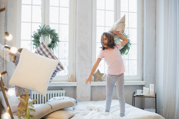 wo Mädchen, Schwestern, die Kissen auf dem Bett kämpfen, das Fenster, das mit einem Weihnachtskranz geschmückt ist, Leben, Kindheit