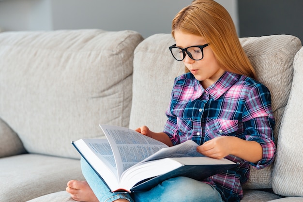 Wo war dieses Kapitel? Schönes kleines Mädchen mit Brille, das Buch liest, während es zu Hause auf der Couch im Lotussitz sitzt