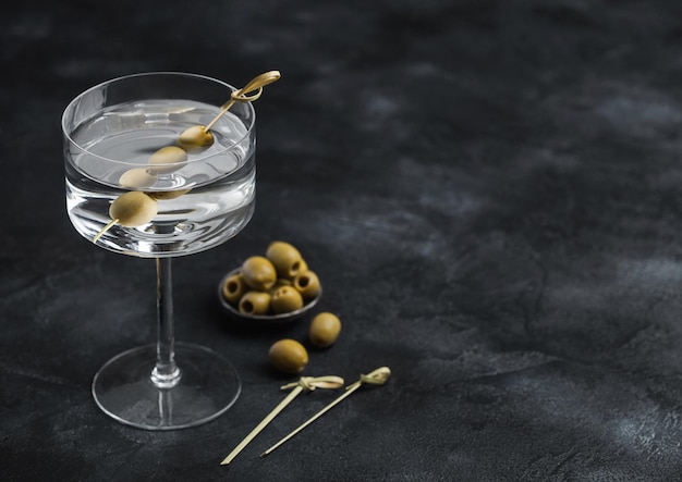 Wodka-Martini-Gin-Cocktail in modernem Glas mit Oliven in Metallschüssel und Bambusstöcken auf Schwarz