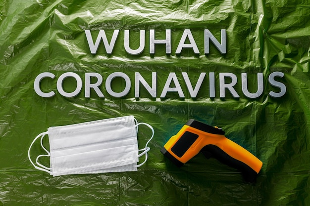 Wörter wuhan coronavirus mit metallbuchstaben auf grünem, zerknittertem plastikfolienhintergrund mit gesichtsmasken und thermometer