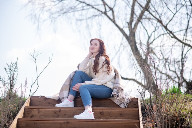 Wohlbefinden, Stimmung. Lächelnde nachdenkliche junge Frau in Pullover und Jeans, die am Frühlingstag auf Treppen in der Natur sitzt sitting