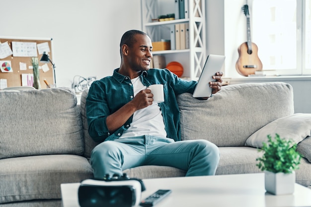 Wohlfühlen. Hübscher junger afrikanischer Mann, der digitale Tablette verwendet und lächelt, während er drinnen sitzt sitting