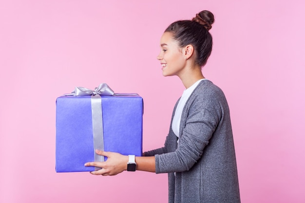 Wohltätigkeitsspende Seitenansicht eines großzügigen jugendlich brünetten Mädchens mit Brötchenfrisur in Freizeitkleidung, das eine Geschenkbox mit freundlichem Lächeln gibt, das Urlaubsgeschenk teilt, Studioaufnahme isoliert auf rosa Hintergrund