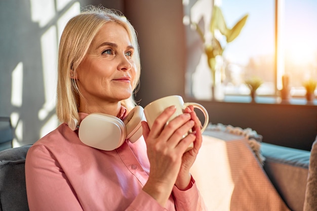 Wohnkomfort und Ruhe Eine schöne ältere Frau hält eine Tasse mit einem Getränk in ihren Händen und lächelt, während sie wegschaut. Kopfhörer um den Hals, Sonnenlicht fällt wunderschön dahinter