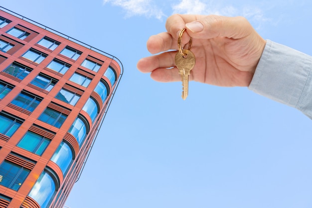 Wohnungsschlüssel in der Hand eines Mannes. Haustürschlüssel aus Messing. Modernes Gebäude, Ansicht von unten. Architektur in der modernen Stadt. Verkauf und Vermietung von Immobilien.