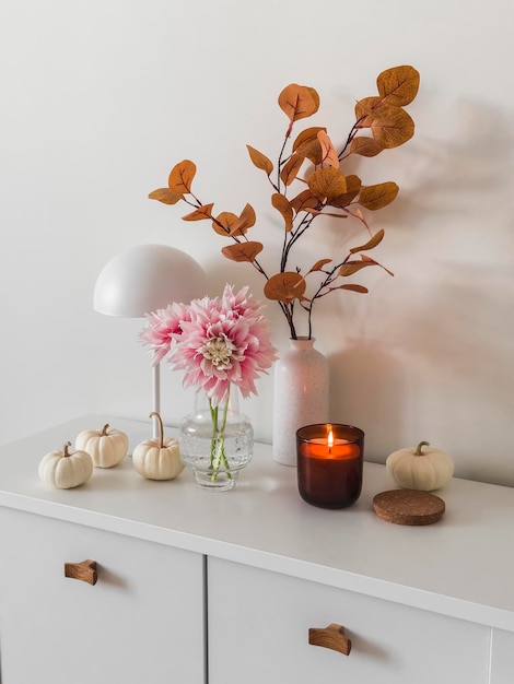 Foto wohnzimmer-interieur im skandinavischen stil, tischlampe, dahlien in einer vase, herbstdekor, brennende kerze auf einer weißen kommode