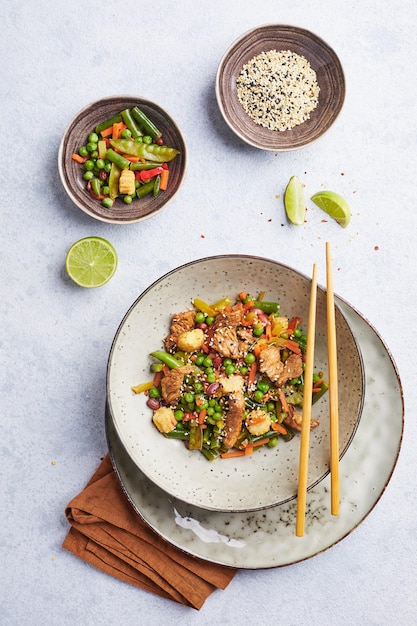 Wok con carne de pavo, maíz, guisantes, judías verdes y zanahorias, servido sobre fondo gris con chopstic