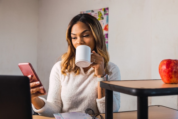 Working at home - mulher latina trabalhando em casa usando seu smartphone e bebendo uma infusão.