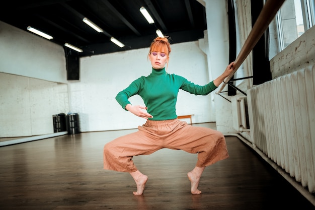 Workout in Bearbeitung. Junger professioneller moderner Tänzer mit rotem Haar, das konzentriert sieht, während er hockt