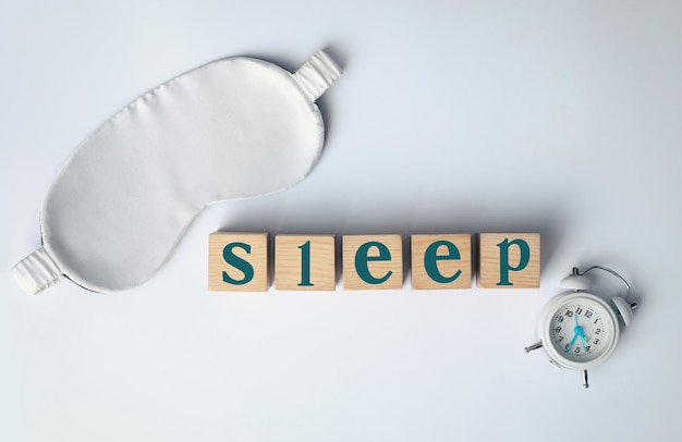 Wort Schlaf aus Holzwürfeln in der Nähe von Augenbinde und Wecker auf weißem Hintergrund flach lag Behandlung von Schlaflosigkeit