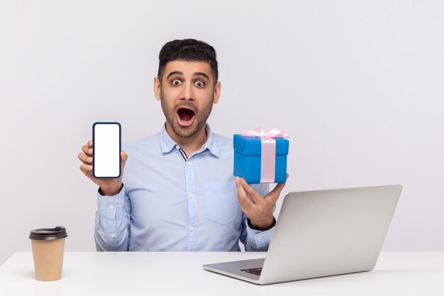 Wow, anuncio de compras en línea. Empleado de oficina asombrado sentado en el lugar de trabajo, sosteniendo una caja de regalo y un teléfono celular con una pantalla en blanco, mirando conmocionado a la cámara. tiro de estudio interior aislado