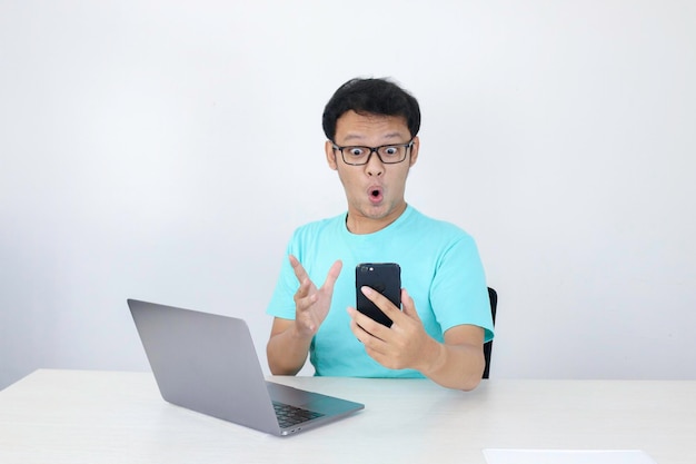 Wow, la cara de un joven asiático se sorprendió por lo que vio en el teléfono con una computadora portátil al lado. Un hombre indonesio con una camisa azul.