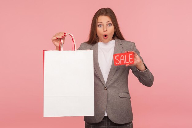 Wow, descuentos en la tienda de moda. Retrato de una mujer de negocios sorprendida y emocionada con chaqueta de traje que parece sorprendida y sostiene la palabra Venta, bolsas de compras con espacio para copiar texto publicitario. foto de estudio