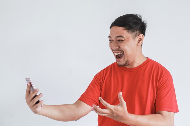 Wow expresión de la cara del hombre asiático feliz con camiseta roja sorprendió lo que ve en el teléfono inteligente