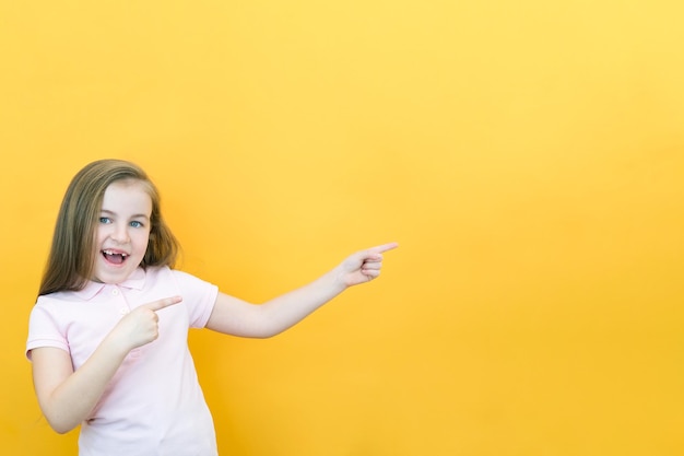 Wow, mira, anuncia aquí Retrato de una linda niña asombrada que señala un espacio vacío en el fondo, una niña preescolar sobre un fondo amarillo